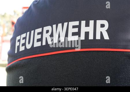 Close-up rear view of a German Feuerwehr firefighter uniform with white lettering and red trim, fire truck blurred in the background during an outdoor Stock Photo
