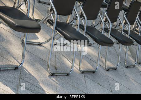 Black folding chairs with chrome frames are arranged in neat rows on a paved outdoor venue, ready for a conference, ceremony, meeting or public event. Stock Photo