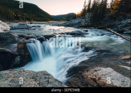 Scenic landscape of long exposure Elbow Falls waterfall rapids flowing over rock formation surrounded by autumn pine forest during sunset in Kananaski Stock Photo