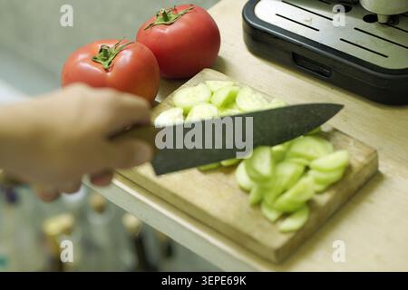 Person's hand holding a knife, preparing fresh vegetables by slicing cucumbers on a wooden chopping board with whole tomatoes nearby, creating a color Stock Photo