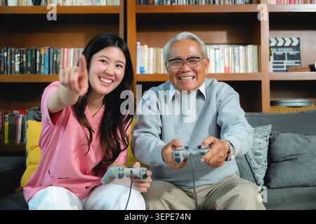 Young woman points toward camera with game controller. Older man smiles while holding controller on couch. Bookshelves with books and film clapboard f Stock Photo