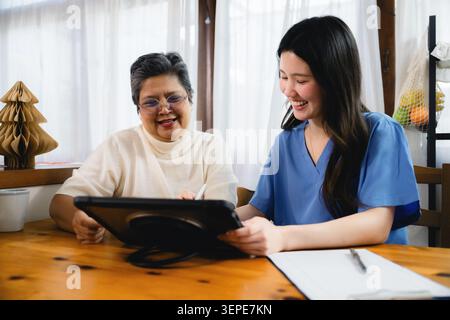Older person sits at wooden table, engaged in conversation. Healthcare worker holds tablet smiling warmly. Discussing healthcare app senior patient ho Stock Photo