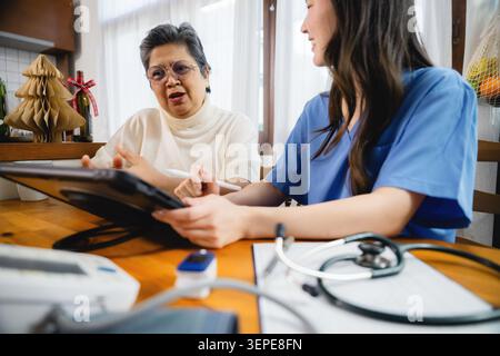Asian grandmother discussing healthcare plan specialist home environment, professional caregiver taking notes tablet, older talking serious and out of Stock Photo
