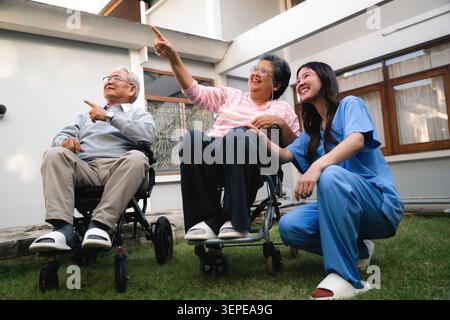 Asian nurse with senior couple wheelchairs pointing finger happy elderly retired husband wife pointing up garden, smiling nurse kneeling beside senior Stock Photo