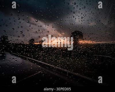Raindrops on a window with a dramatic sunset glowing over a dark landscape during an evening storm Stock Photo