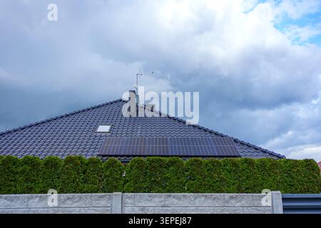 A modern residential house featuring a dark grey tiled roof equipped with photovoltaic solar panels, partially hidden behind a green hedge and a concr Stock Photo
