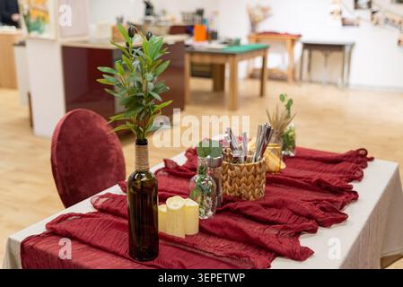 Dining table decorated with rustic olive branch in glass bottle, cutlery basket, and candles on red textured fabric for stylish event settings. Stock Photo