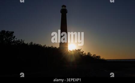 A tall lighthouse stands in silhouette against a darkening evening sky, as the setting sun bursts through a line of trees at its base. Stock Photo