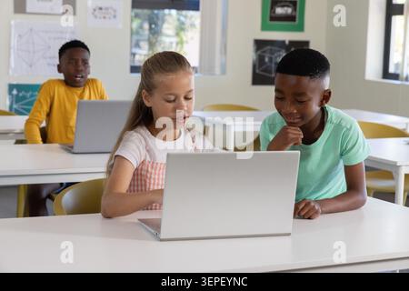 Diverse child classmates collaborating on silver laptop in bright classroom, girl in pink pinafore Stock Photo