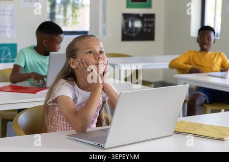School-age classmates sitting at desks in bright classroom, girl daydreaming and boys using laptops Stock Photo