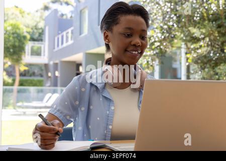 Youth African female sitting on patio, using laptop, writing in notebook, wearing pink headphones Stock Photo