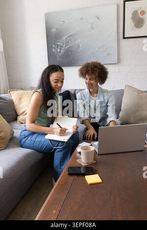 African female friends sitting on gray couch in living room, writing in notebook and using laptop Stock Photo