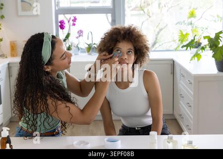 African American women applying blue gel eye patches at kitchen countertop with skincare bottles Stock Photo