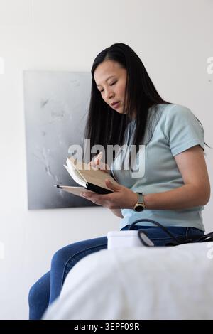 Mature Asian woman sitting on bed, writing in notebook with pen, wearing blue top Stock Photo