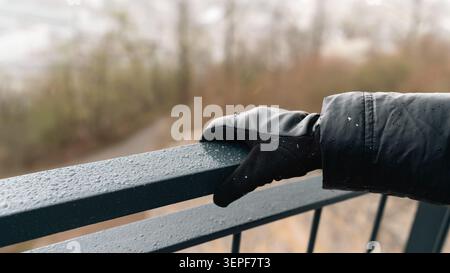 Close-up of a hand in a black sleeve resting on a wet metal railing covered with raindrops, creating a cold outdoor weather detail Stock Photo