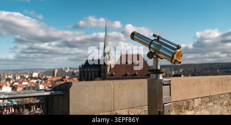 Erfurt, Germany - March 27, 2026. Viewing telescope beside locks on Petersberg with blurred Erfurt Cathedral, rooftops, blue sky, and clouds in the ba Stock Photo