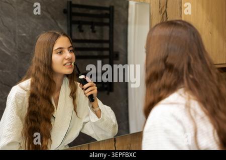 Young woman brushing teeth with electric toothbrush. Young woman in a white waffle bathrobe brushing her teeth with an electric toothbrush while Stock Photo