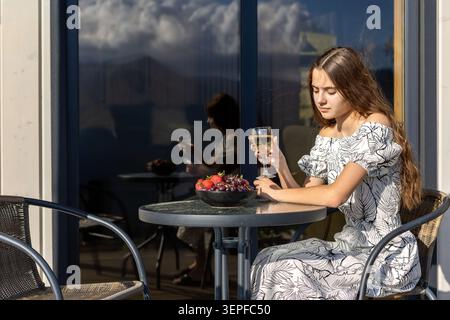 Young woman with fruit and drink on sunny balcony. Young woman sits at a sunny balcony table holding a glass beside a bowl of strawberries and grapes. Stock Photo