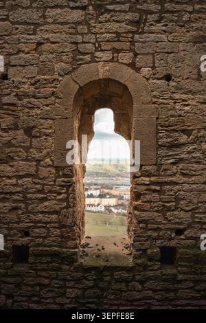 Historic stone castle window framing a distant European town, green valley, and forested hills, with textured medieval masonry in the foreground Stock Photo