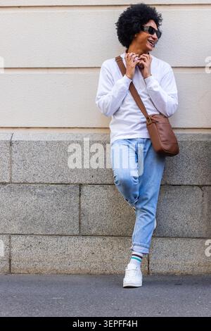 African American man leaning on granite ledge on sidewalk by beige wall, smiling with shades, bag Stock Photo