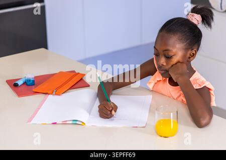 African child writing in open notebook on kitchen counter with green pencil, peach top, copy space Stock Photo