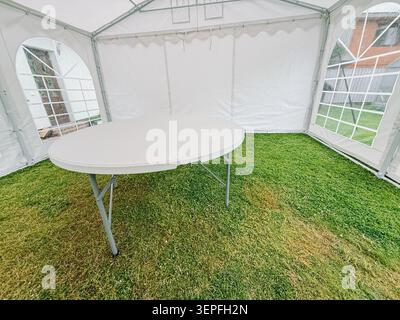 Smiltene, Latvia - June 12, 2026: Close-up view of a round folding table inside a white marquee tent with a natural grass floor. Clean and versatile e Stock Photo
