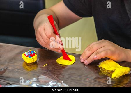 A child is using a red tool to shape yellow modeling clay on a table. Small pieces of clay are nearby. The child focuses on creating a design. The set Stock Photo