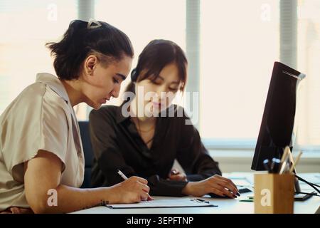 Young adult businesswomen working in call center office, reviewing documents and computer data during team support tasks. Ideal for customer service, teamwork, training marketing Stock Photo