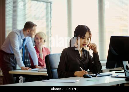 Asian young adult woman wearing headset, taking customer support call and noting details at office desk while team colleagues collaborating behind. Ideal for customer service, CRM, outsourcing Stock Photo