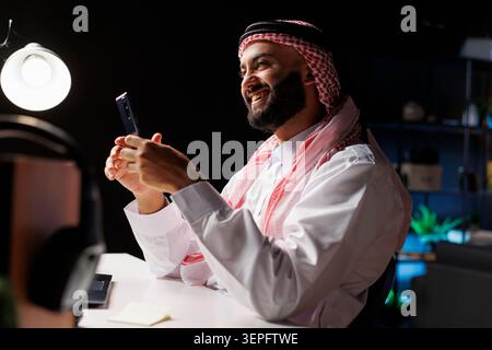 Side-view of smiling Muslim man in professional attire works at a modern desk. Using a smartphone, he browses the internet, representing the digital age and wireless technology. Stock Photo