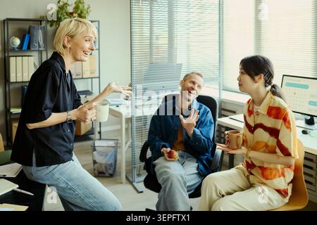 Diverse young adult coworkers chatting during office break, holding mugs and discussing ideas in workplace. Ideal for teamwork, company culture, collaboration, office communication Stock Photo