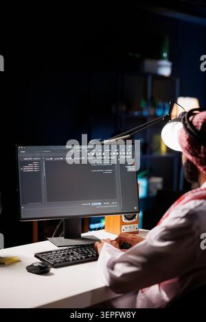 Traditional Middle Eastern developer compiles code on desktop computer, showcasing expertise in programming, algorithms, and software development; selective focus on pc monitor displaying source code. Stock Photo