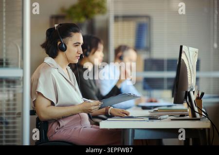 Young woman with disability in wheelchair wearing headset, reviewing notes and assisting customers in call center office with team support. Ideal for customer service, CRM, outsourcing marketing Stock Photo