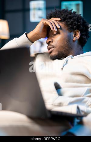 Teleworker having health problems, suffering from headache and nausea, sitting on couch at home, trying to work; remote employee suffering from illness, struggling to input data on laptop. Stock Photo