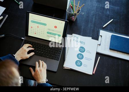 High angle view of young male employee using laptop and reviewing business calendar on desk, managing schedule and productivity. Suitable for remote work, planning, finance marketing Stock Photo