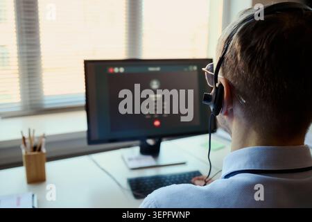 Rear view of young Caucasian man wearing headset, handling customer support call at office workstation. Ideal for call center, client service, help desk marketing Stock Photo