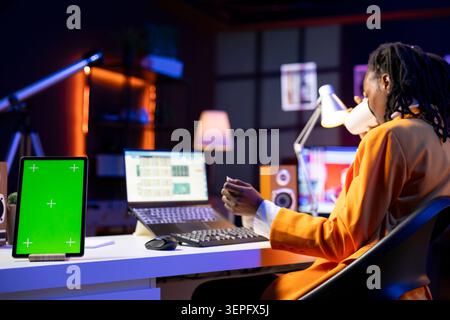 Teleworker in home office completing spreadsheets on laptop next to mockup tablet, entering data, calculating totals; green screen device next to bookkeeper using spreadsheet software. Stock Photo