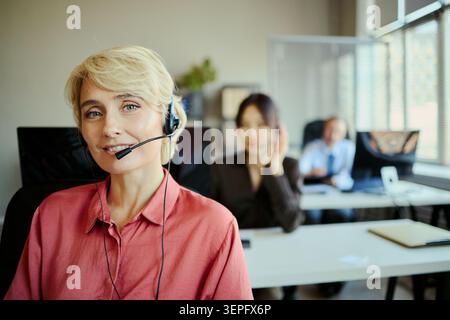Portrait of smiling Caucasian young adult woman wearing headset and assisting customers in call center office, representing client support and team service for business marketing Stock Photo
