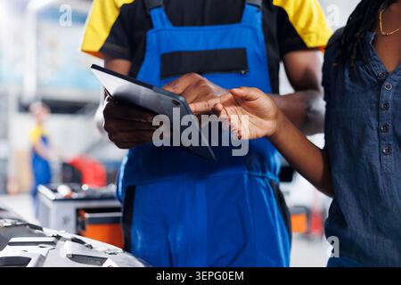 Mechanic in car service uses laptop to calculate invoice for client after repairing brakes while BIPOC employee in garage with customer calculating final costs after servicing vehicle. Stock Photo