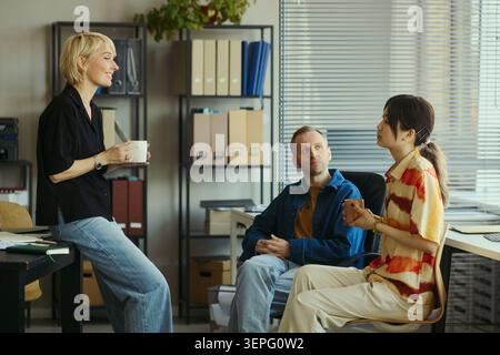 Diverse young adult coworkers discussing project strategy during office meeting, holding coffee and collaborating on decisions. Ideal for teamwork, business planning, workplace communication Stock Photo
