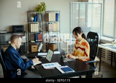 Diverse young adult coworkers collaborating at office desk, typing on laptops and taking notes during meeting, representing teamwork and productivity. For business planning or startup marketing Stock Photo