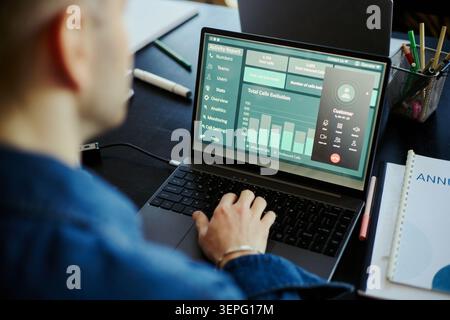Young male office worker monitoring call center analytics on laptop, managing customer support workflow and team performance. Ideal for customer service, CRM, KPI, remote support Stock Photo