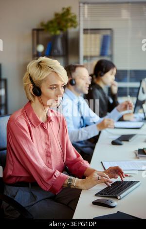 Vertical shot of Caucasian young adult woman wearing headset, typing at call center desk while team assisting clients in office. Ideal for customer support, CRM, outsourcing marketing Stock Photo