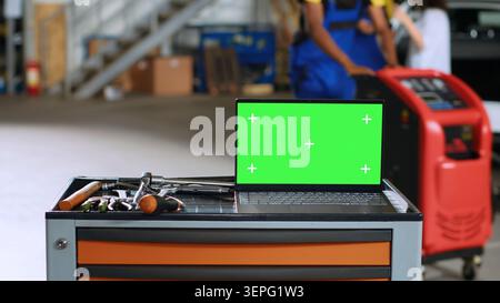 Green screen laptop placed on working bench in busy garage next to professional tools while mechanics walk around in blurry background. Chroma key device in auto repair shop, close up. Stock Photo