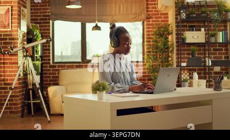 Freelancer enjoys working at workstation while listening to music and performing internet activities, at sunset, African American woman is multitasking, online career in blogging. Stock Photo