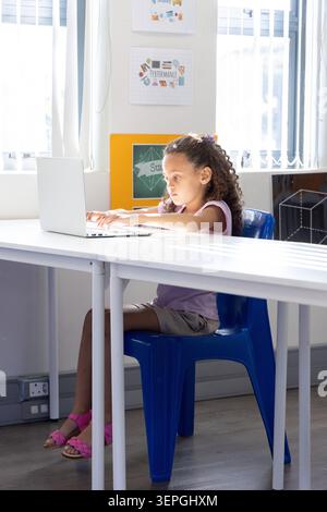 Female child sitting on blue plastic chair using silver laptop at classroom table with posters Stock Photo