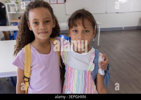 Two school-age girls standing together in bright classroom, wearing backpacks near whiteboard Stock Photo
