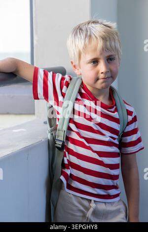 Blond boy child leaning on concrete railing at stair landing, wearing striped t-shirt and backpack Stock Photo