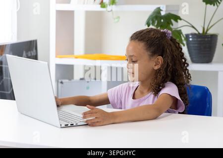 Female child sitting in blue plastic chair, typing on silver laptop at white desk with shelving Stock Photo