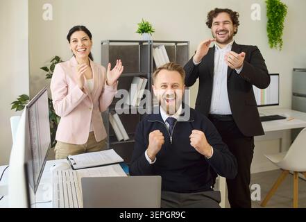 Excited office team celebrating business success with cheering colleague by laptop Stock Photo